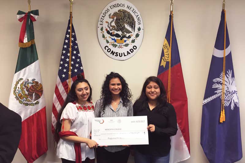 Three Meredith students in front of Mexican, U.S., N.C. and S.C. flags, holding a presentation check representing scholarship funds