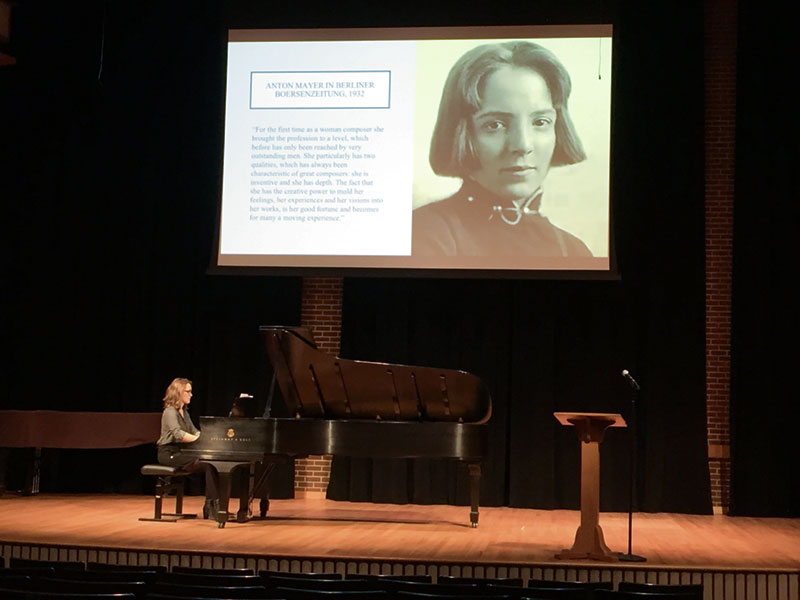 Student plays piano during a presentation about composer Sophie Eckhardt-Gramatte, whose photo is shown on a screen above the piano 