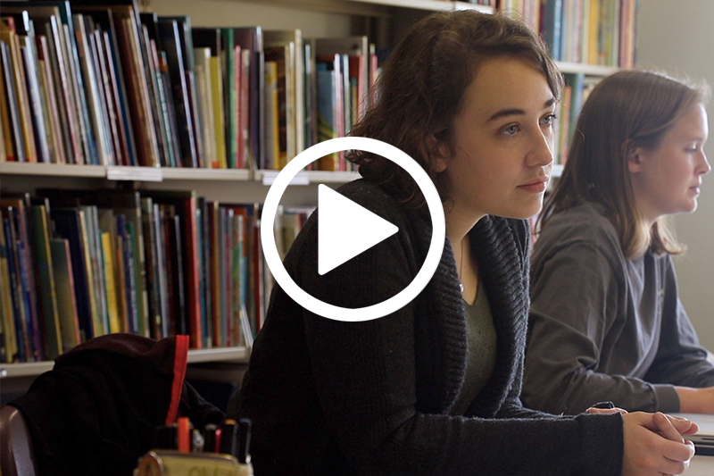 Click on image of students seated at desk in front of library book stack to watch video in modal