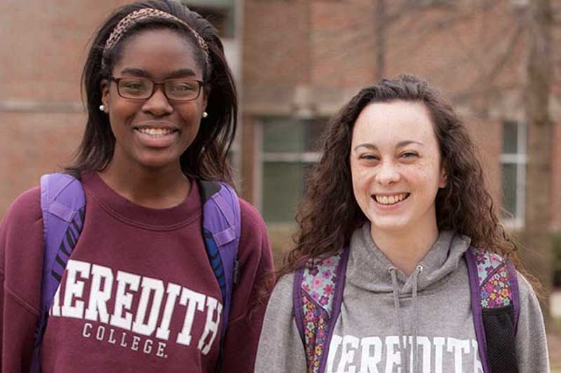 Two women students wearing Meredith shirts standing outside a classroom building