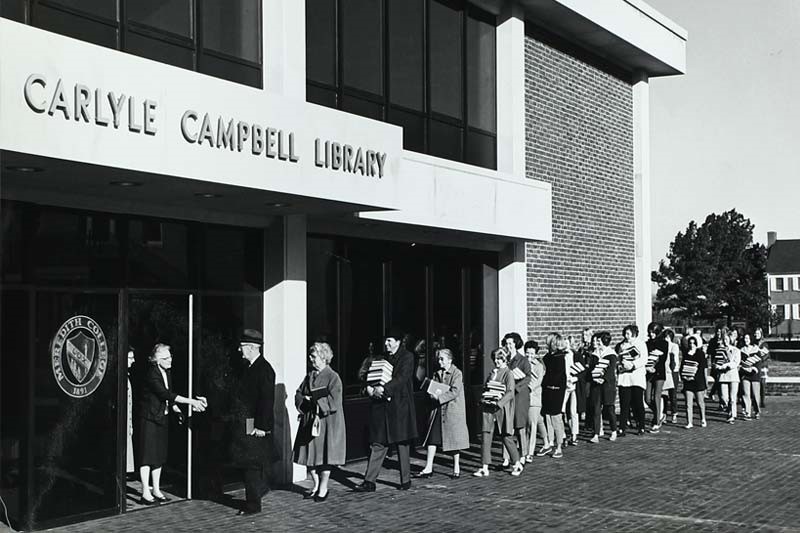 Photo shows the 1969 Book In, during which students helped move books to the new location. The photo shows a line of people holding books waiting to go into the building