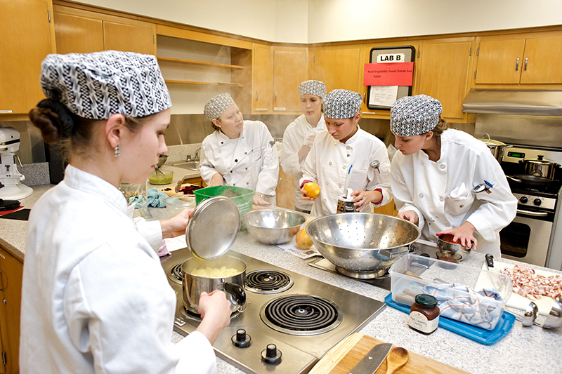 Students in white chef robes prepare a meal in a kitchen at Meredith’s nutrition program.