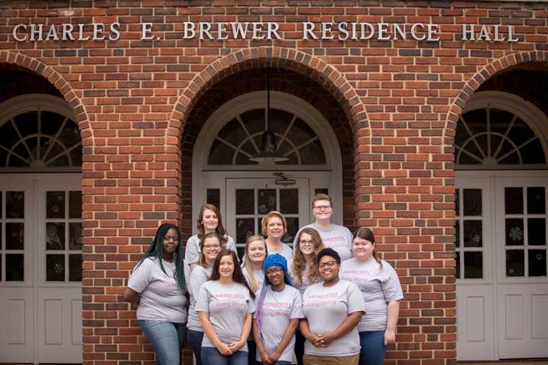 Ten students and one teacher taking a group photo in front of Brewer Residence Hall