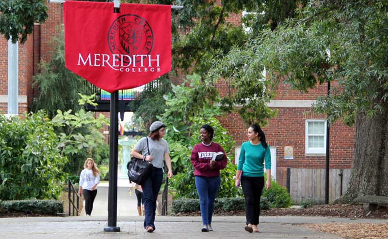 Three students walk together on Meredith's campus. A Meredith College banner, the words Meredith College in white on a maroon background, is in the foreground of the photo.