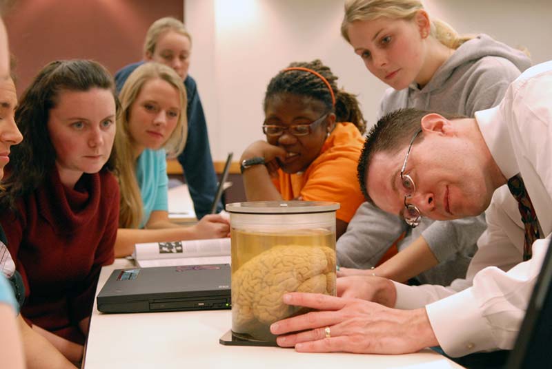 Professor of Psychology Mark O'Dekirk with a group of students gathered around as he shows a brain in a class jar.