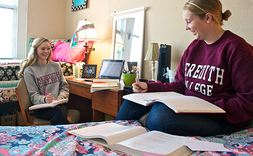 Two women students wearing Meredith shirts sit together in their residence hall room.