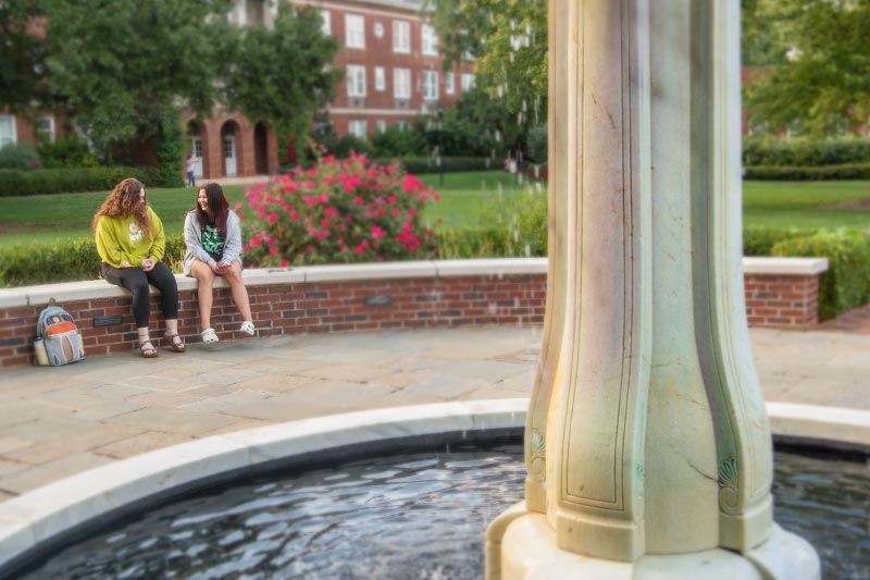 Two students sitting on brick wall at fountain in courtyard.