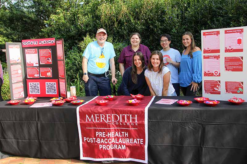 Provost Matthew Poslusny, holding a cane in his left hand, with a group of students at the donate plasma table set up by the pre-health post-baccalaureate program.