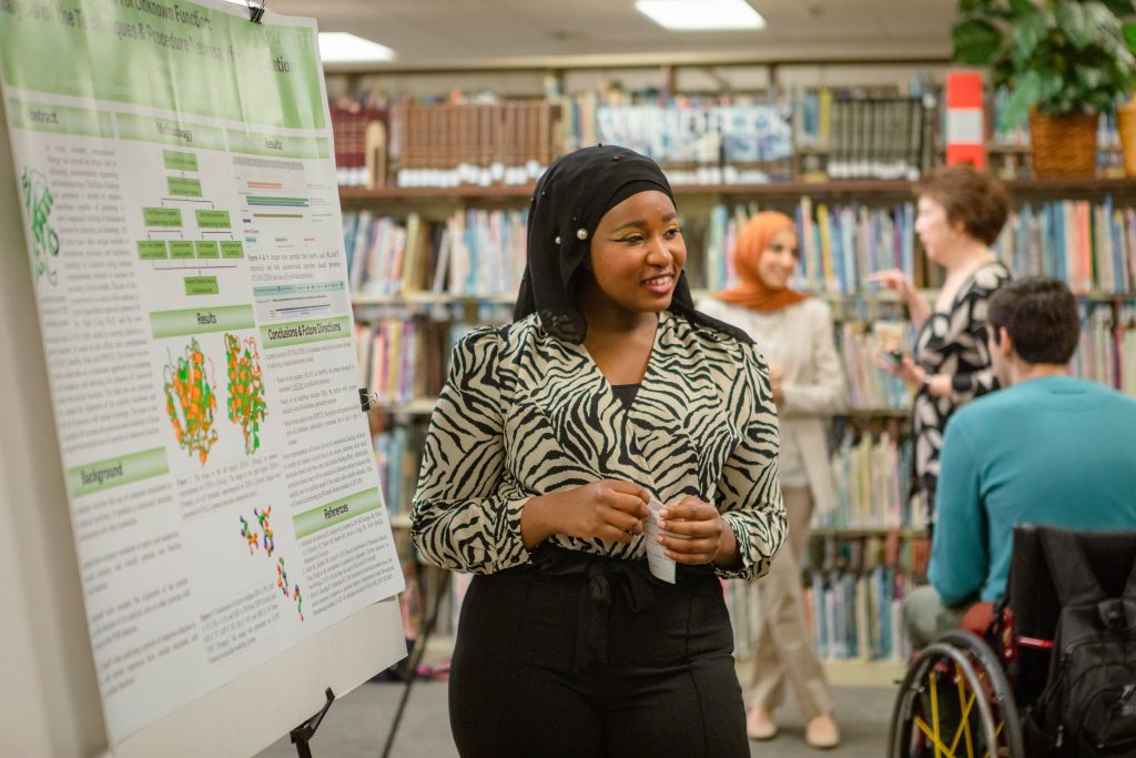 A student wearing a head covering smiles as she presents her research at a poster presentation.